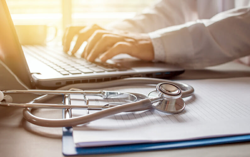 Clinician's hands type on a laptop with a stethoscope in the foreground